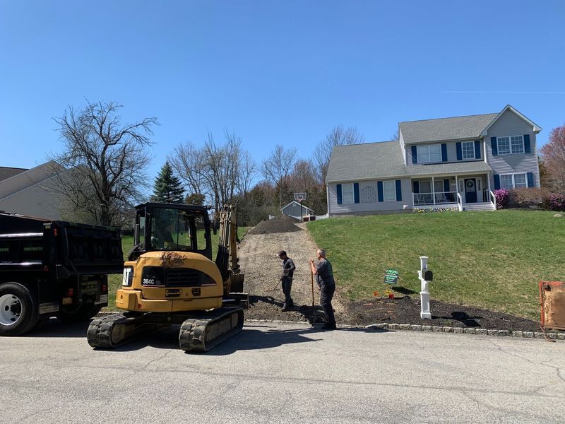 Construction crew paving a driveway in front of a house. Excavator and dump truck present. Sunny day.