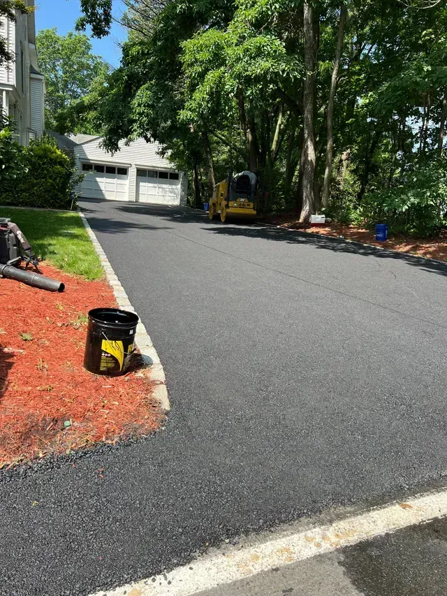 Newly paved asphalt driveway with a compacting machine, bordering red mulch and lawn.