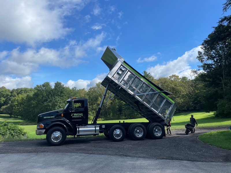 Black dump truck with raised bed on driveway, green trees in background, sunny sky.