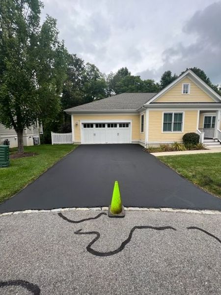 Freshly paved asphalt driveway leading to a yellow house with a two-car garage. A traffic cone sits on the driveway.