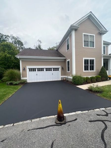 Newly paved black driveway in front of a tan two-story house with a white garage door.