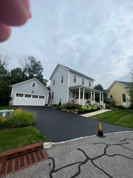 Two-story gray house with detached garage and black driveway. Green lawn, flowers, and cloudy sky.