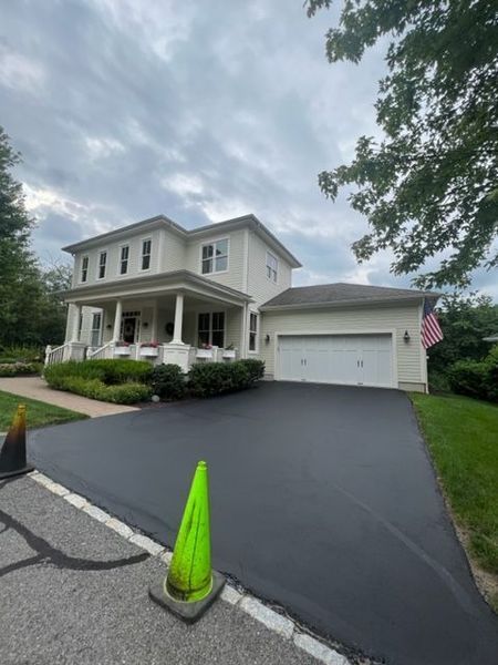 Two-story house with a newly paved black driveway. A green cone is in the foreground. Cloudy sky.