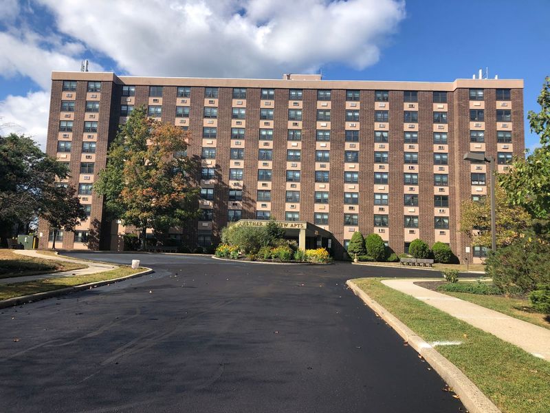 A tall, brown brick apartment building on a sunny day with blue sky and a newly paved driveway.