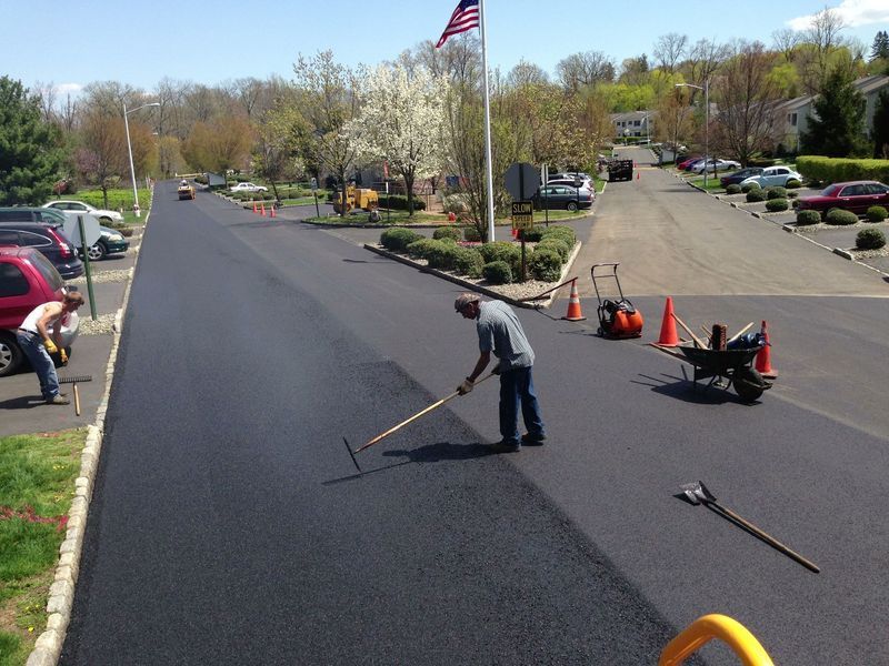 Road workers paving a street; one rakes asphalt, others work nearby; sunny day, flag in the background.
