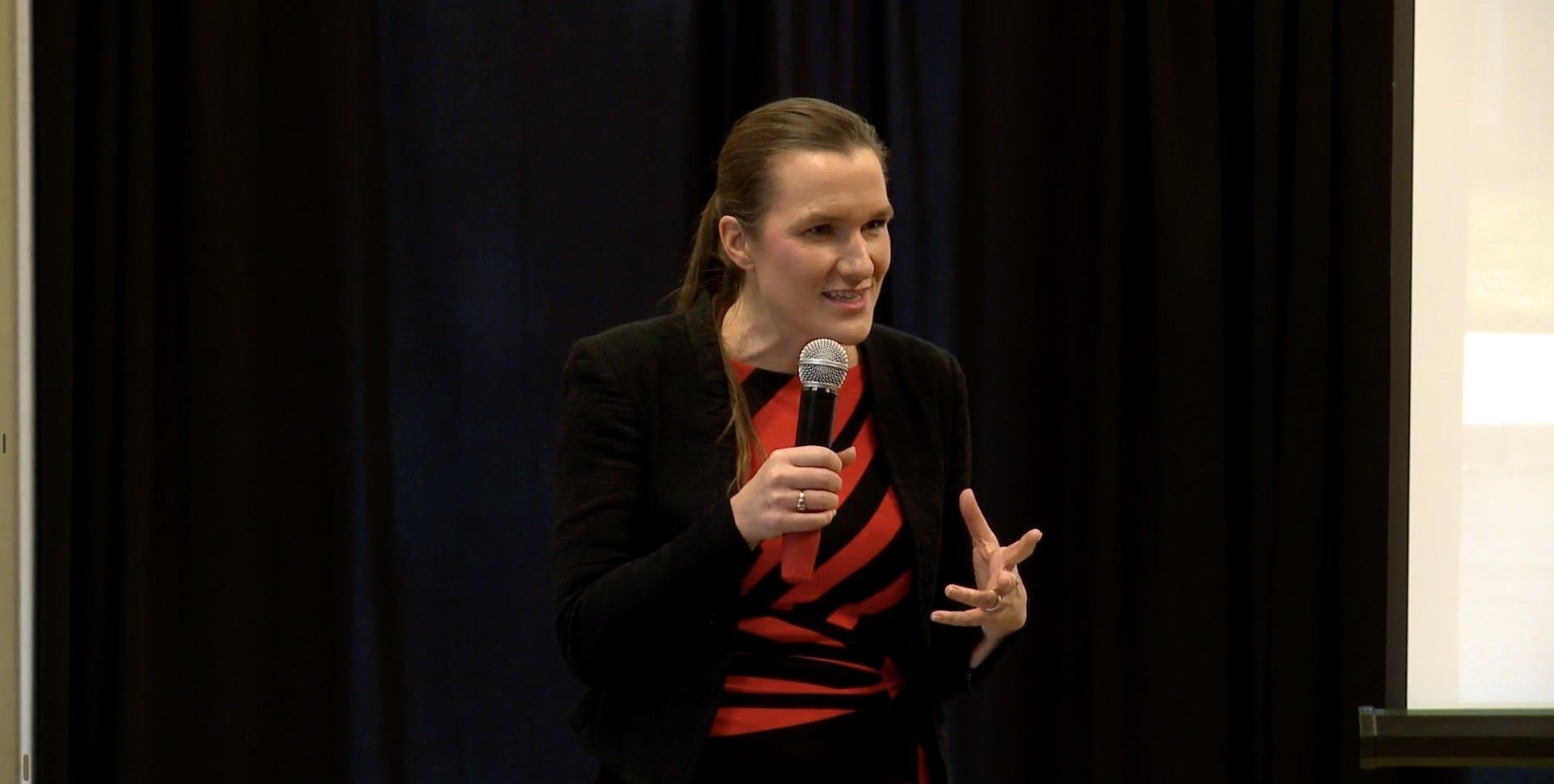 Woman speaking into a microphone on stage, wearing a black jacket and red striped shirt.