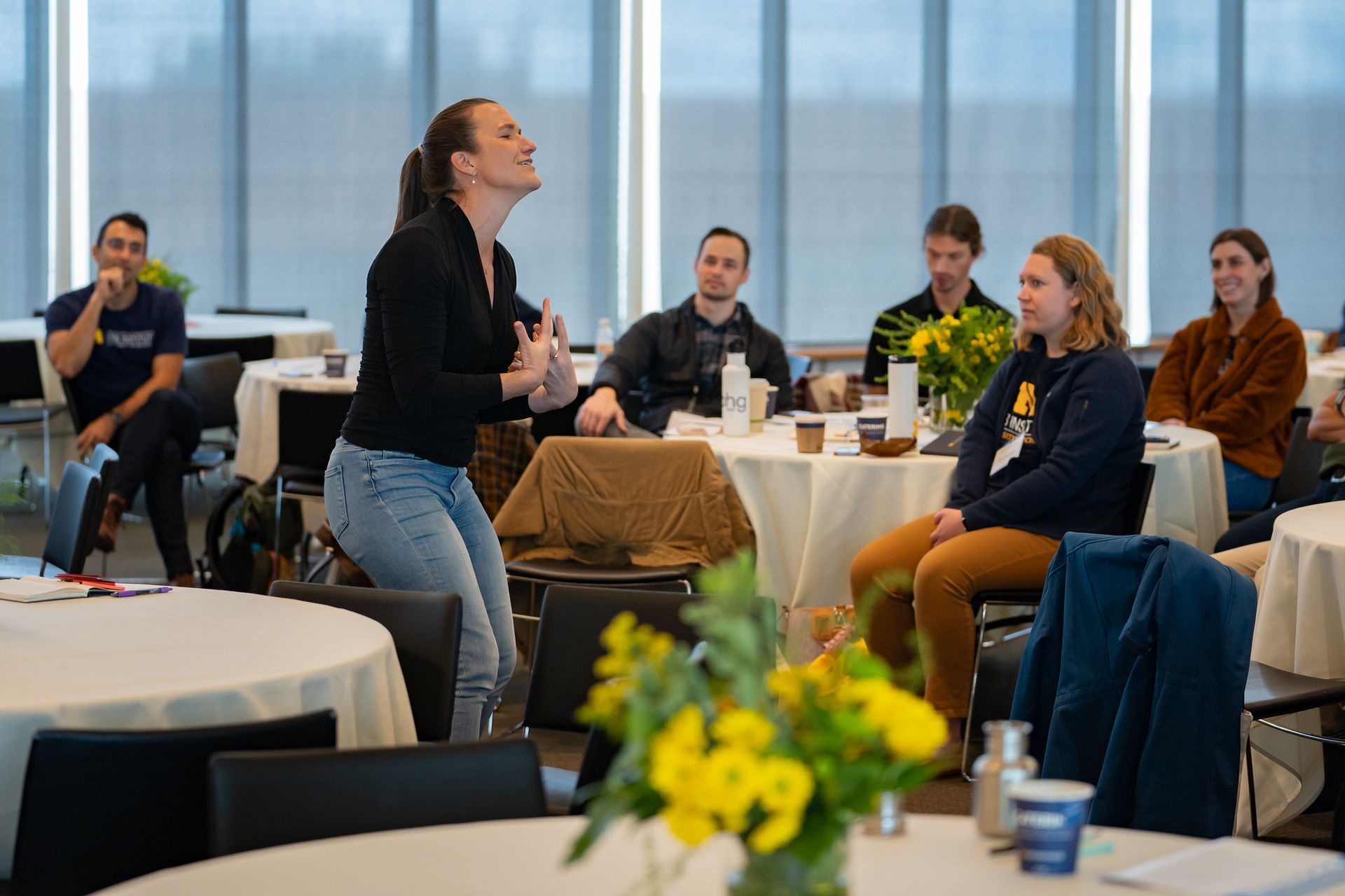 Woman speaking to seated audience in a bright conference room, with tables and a yellow flower arrangement.