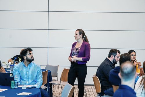 People gathered at round tables in a conference room; a woman in a purple top stands speaking.