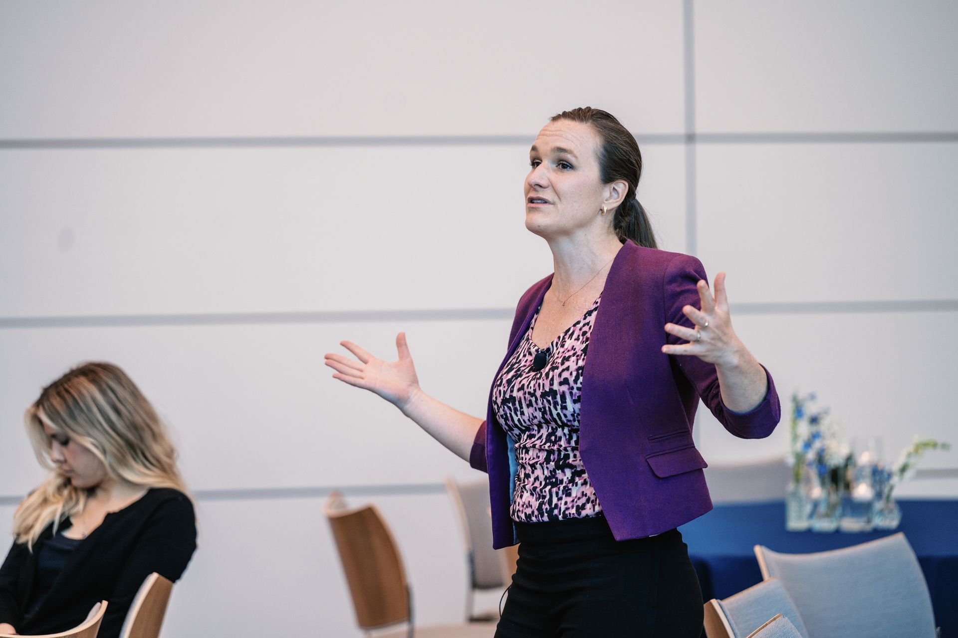 Woman speaking at a conference, wearing a purple jacket and gesturing on stage while another woman sits nearby.