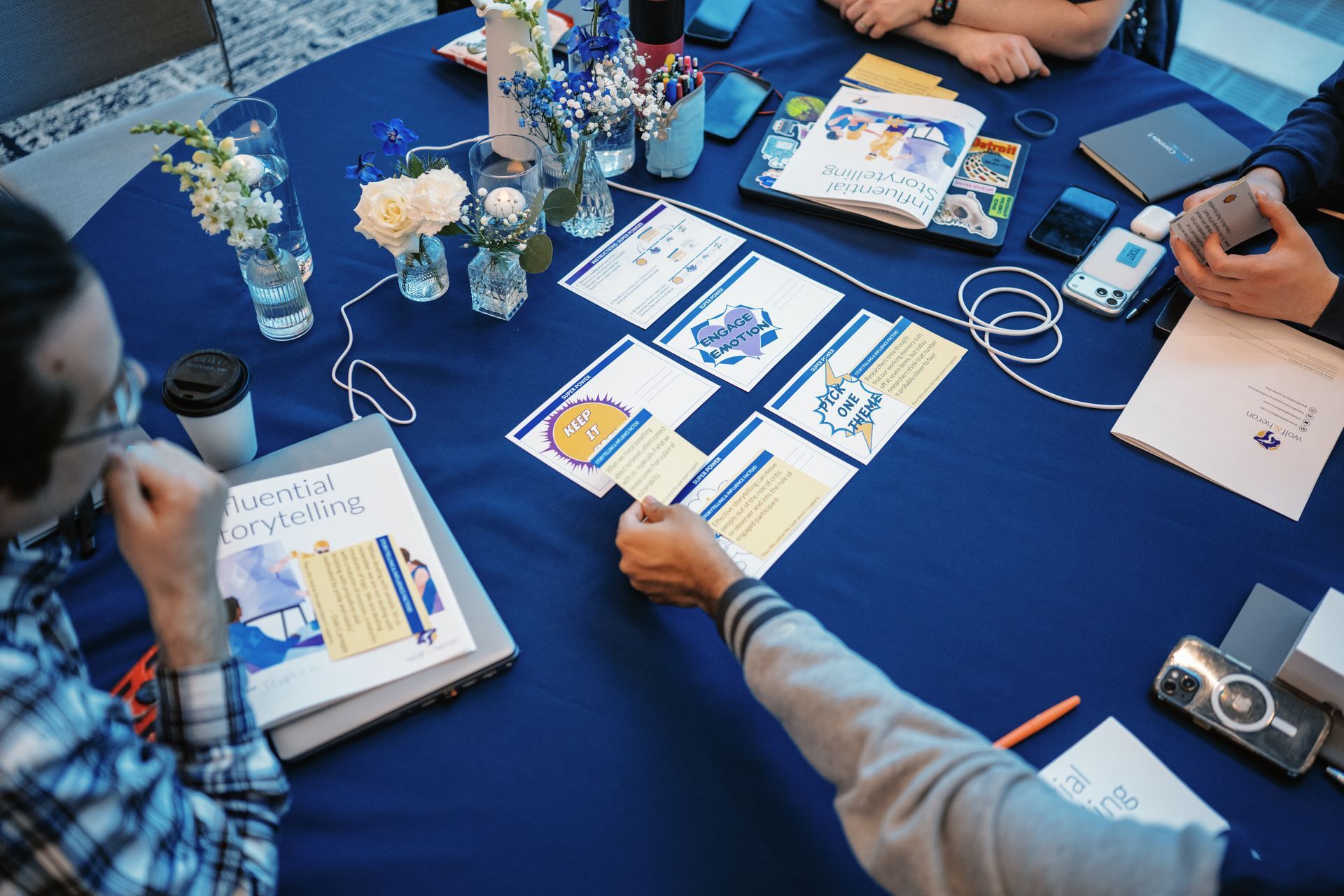 People reviewing printed charts and notes around a blue table with flowers and cameras