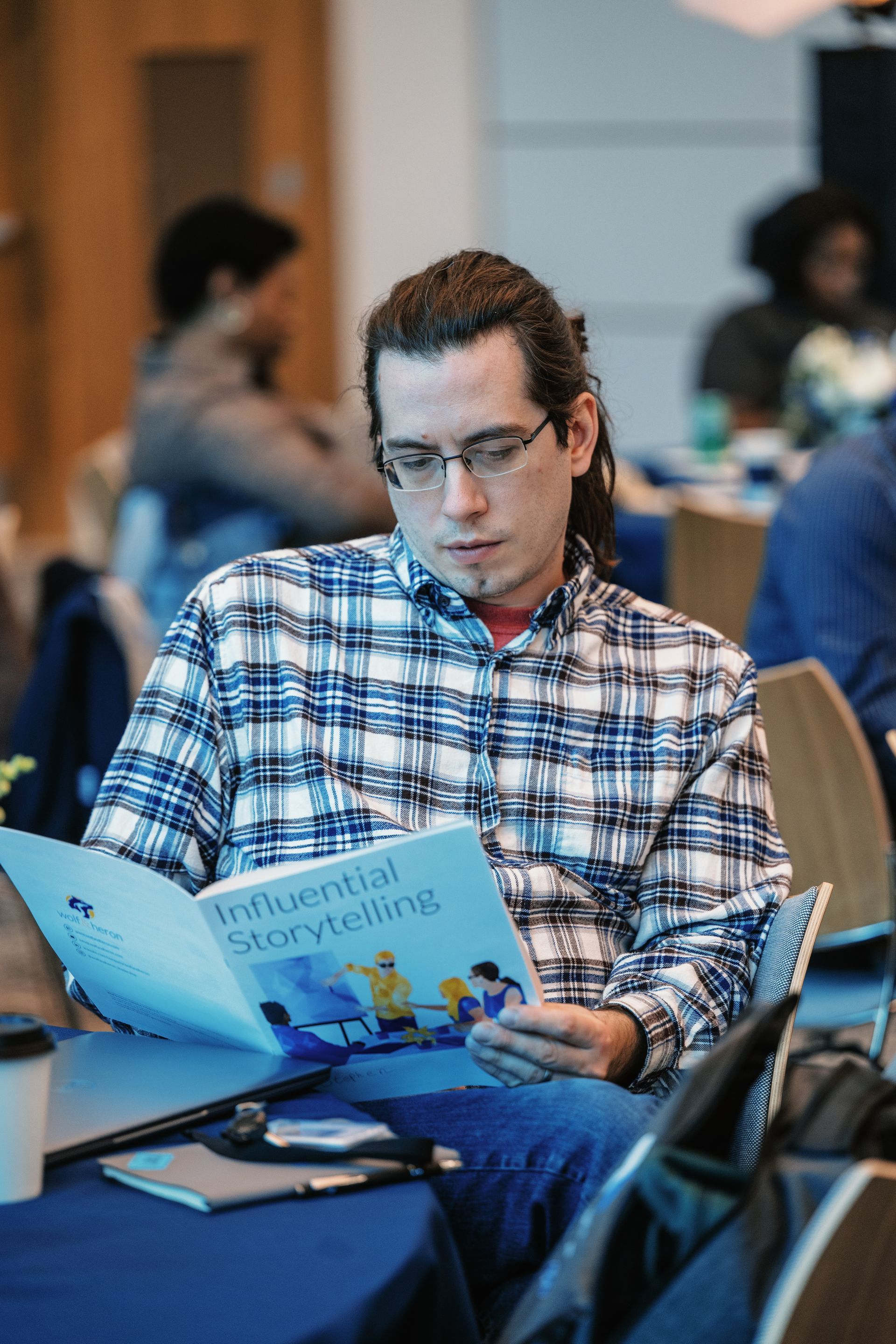 Man in a blue plaid shirt reading a magazine at a table in a café or event space