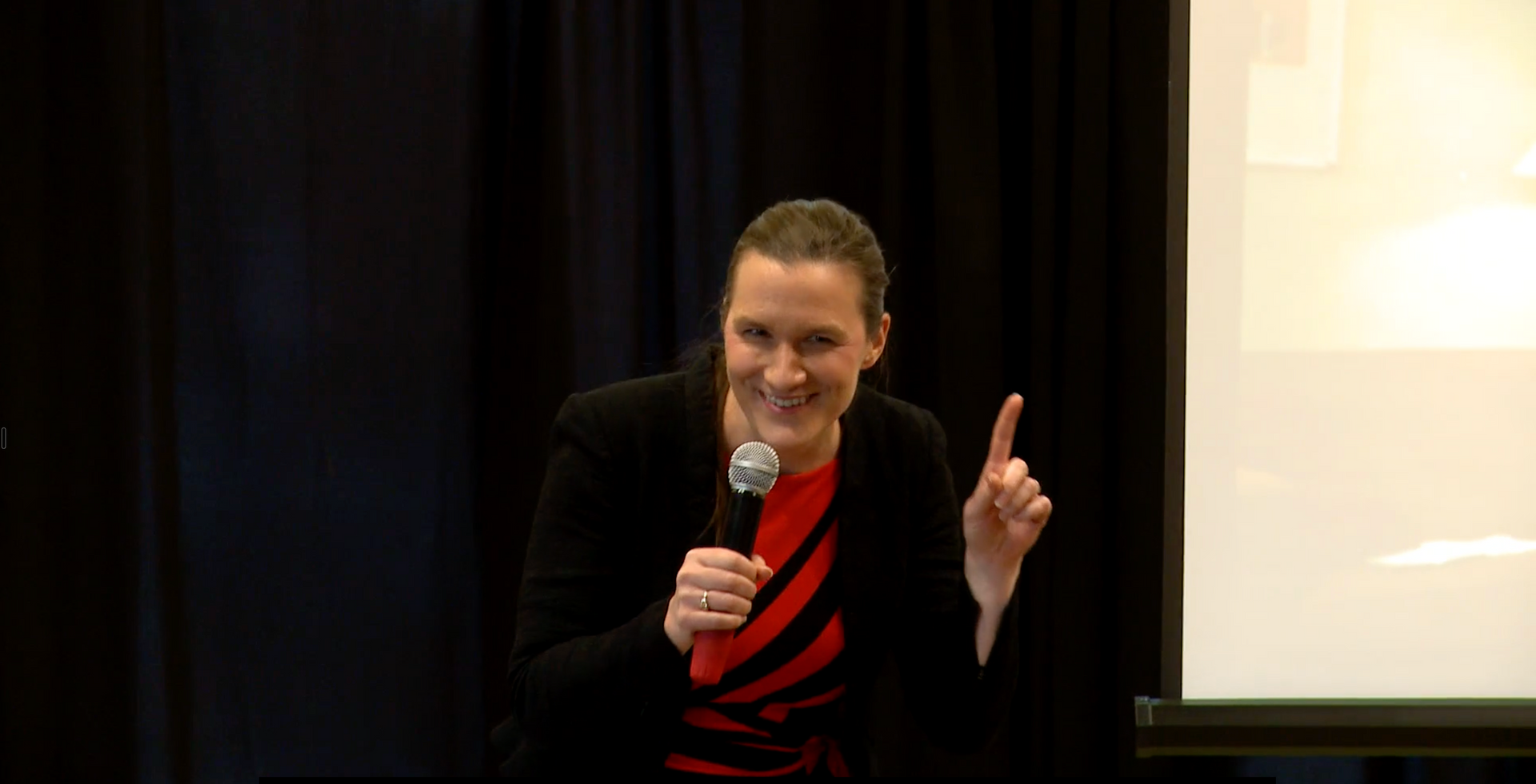 Smiling speaker holding a microphone and pointing upward on a stage beside a projection screen