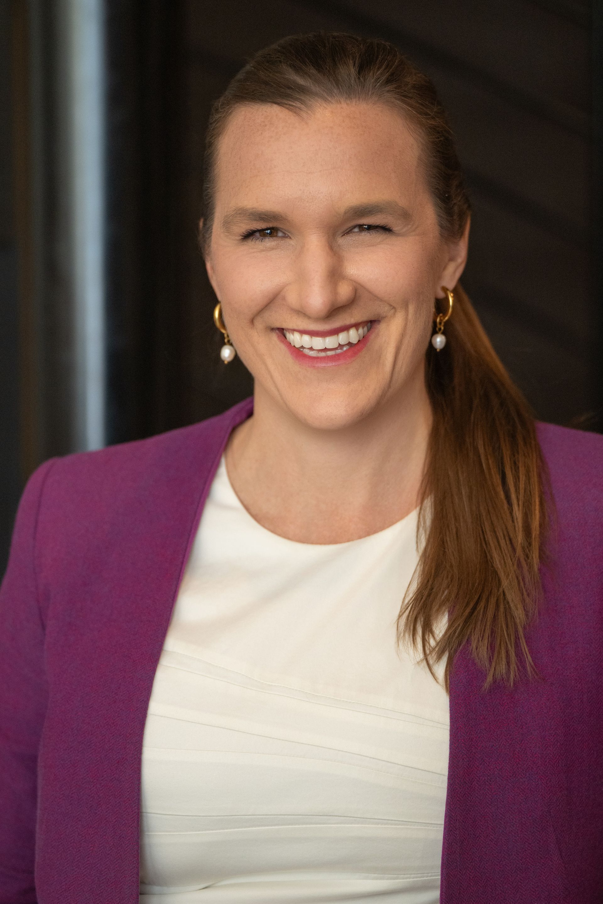 Smiling person in a purple blazer and white top against a dark background