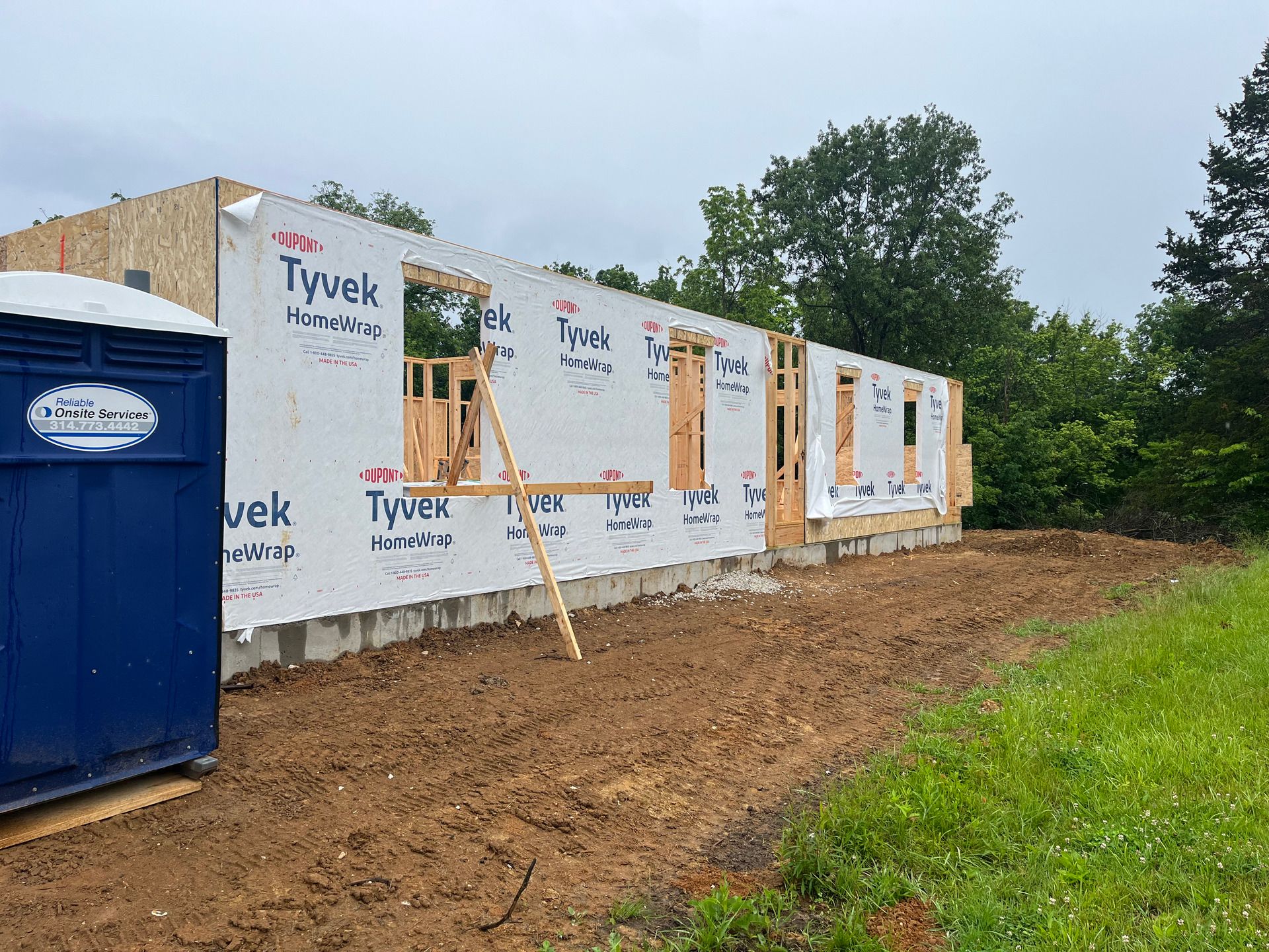House under construction wrapped in Tyvek, with blue portable toilet and dirt lot in front