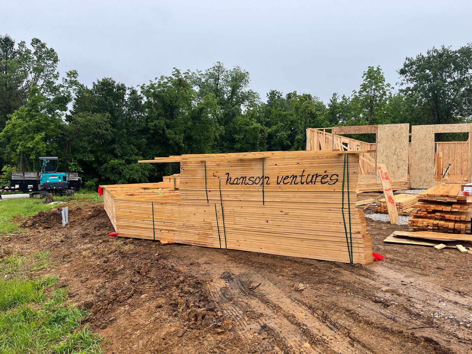 Stacks of lumber and plywood at a muddy construction site with a truck and trees in the background