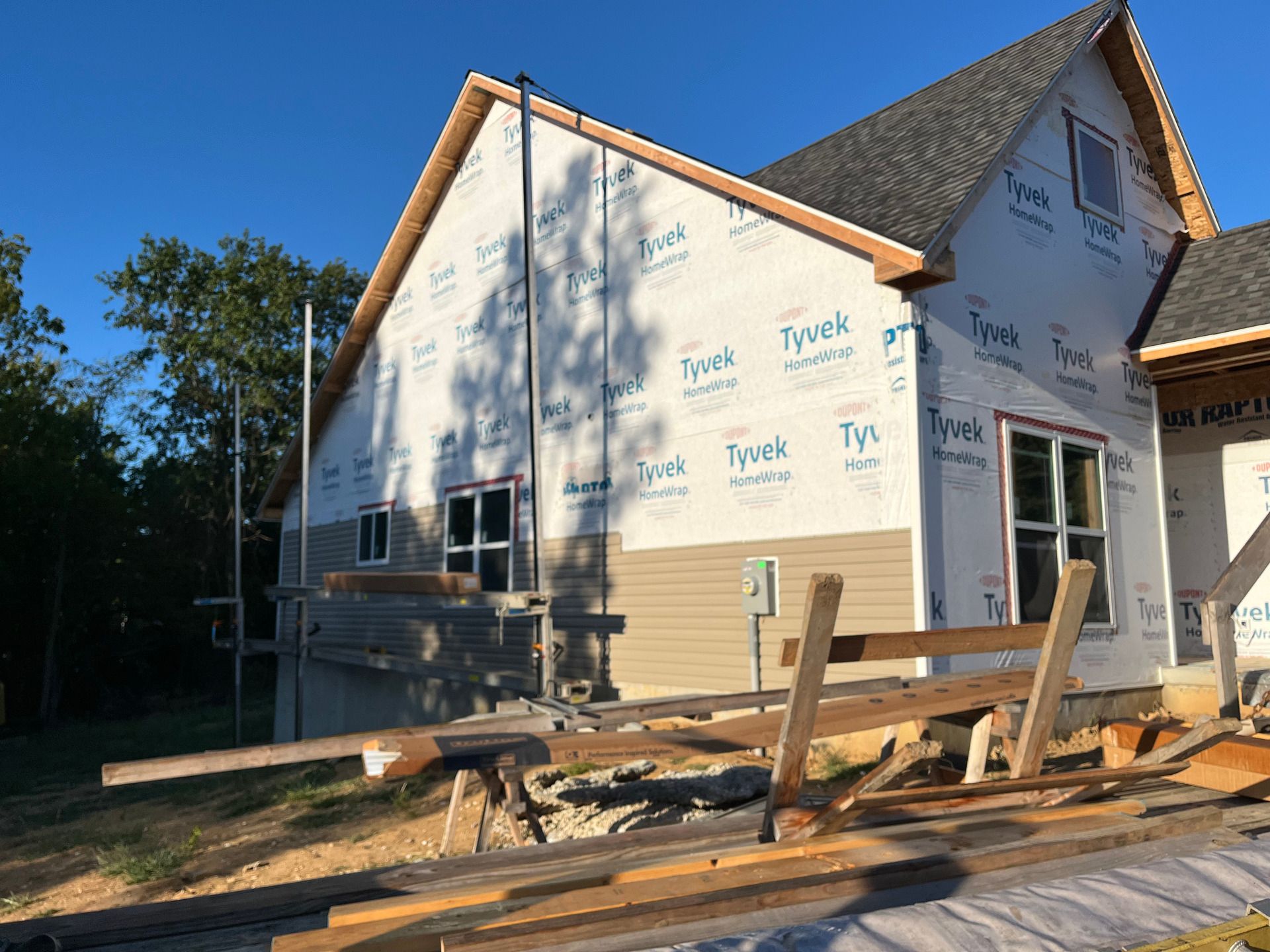 Partially built house with white Tyvek wrap, exposed wood framing, and construction debris in the yard