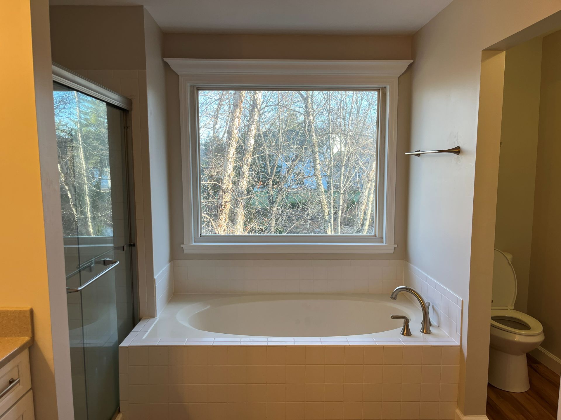 Bathroom with a white soaking tub beneath a large window and beige walls, overlooking trees outside