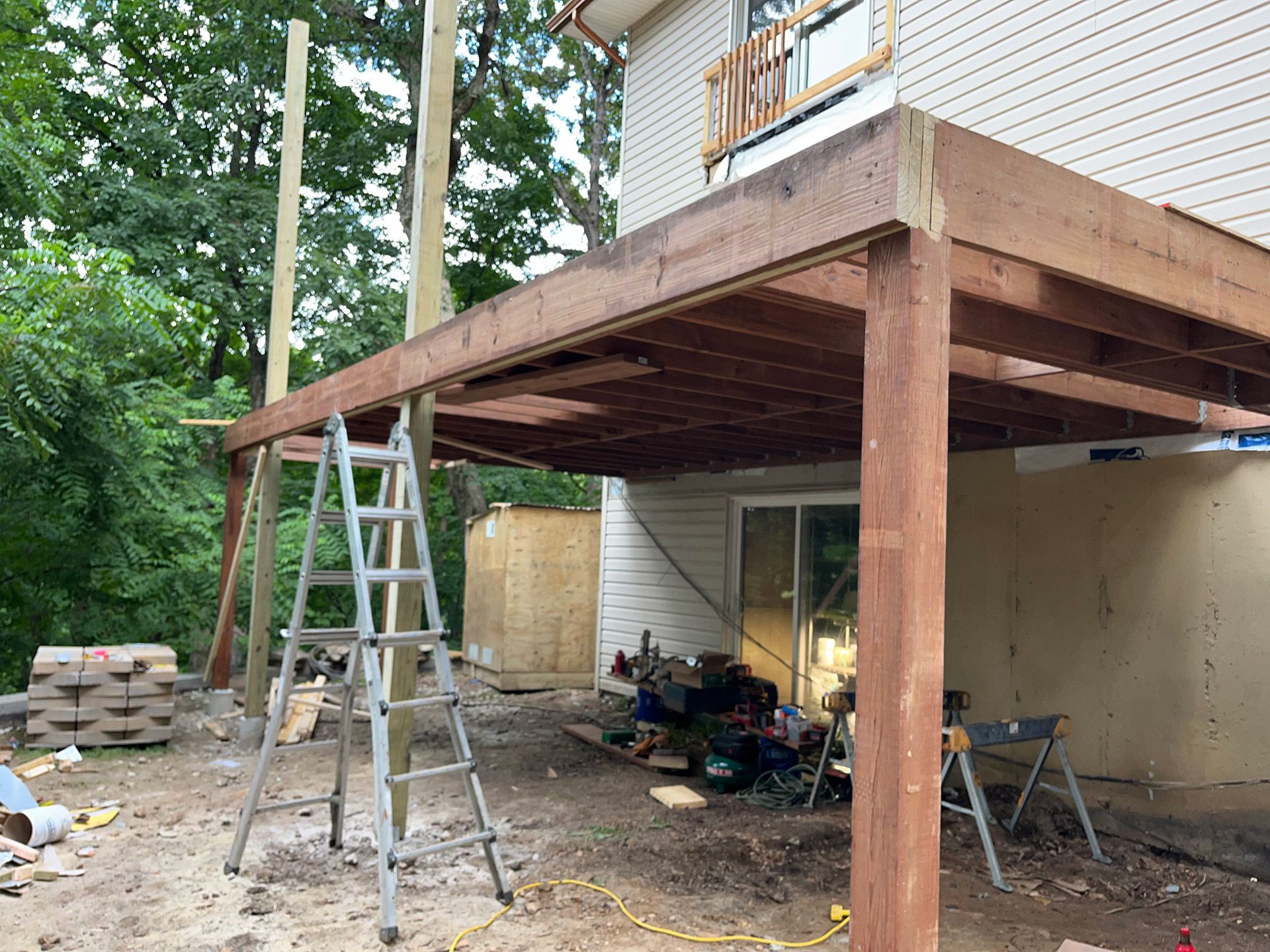 Wooden deck under construction beside a house, with ladders and building materials underneath.