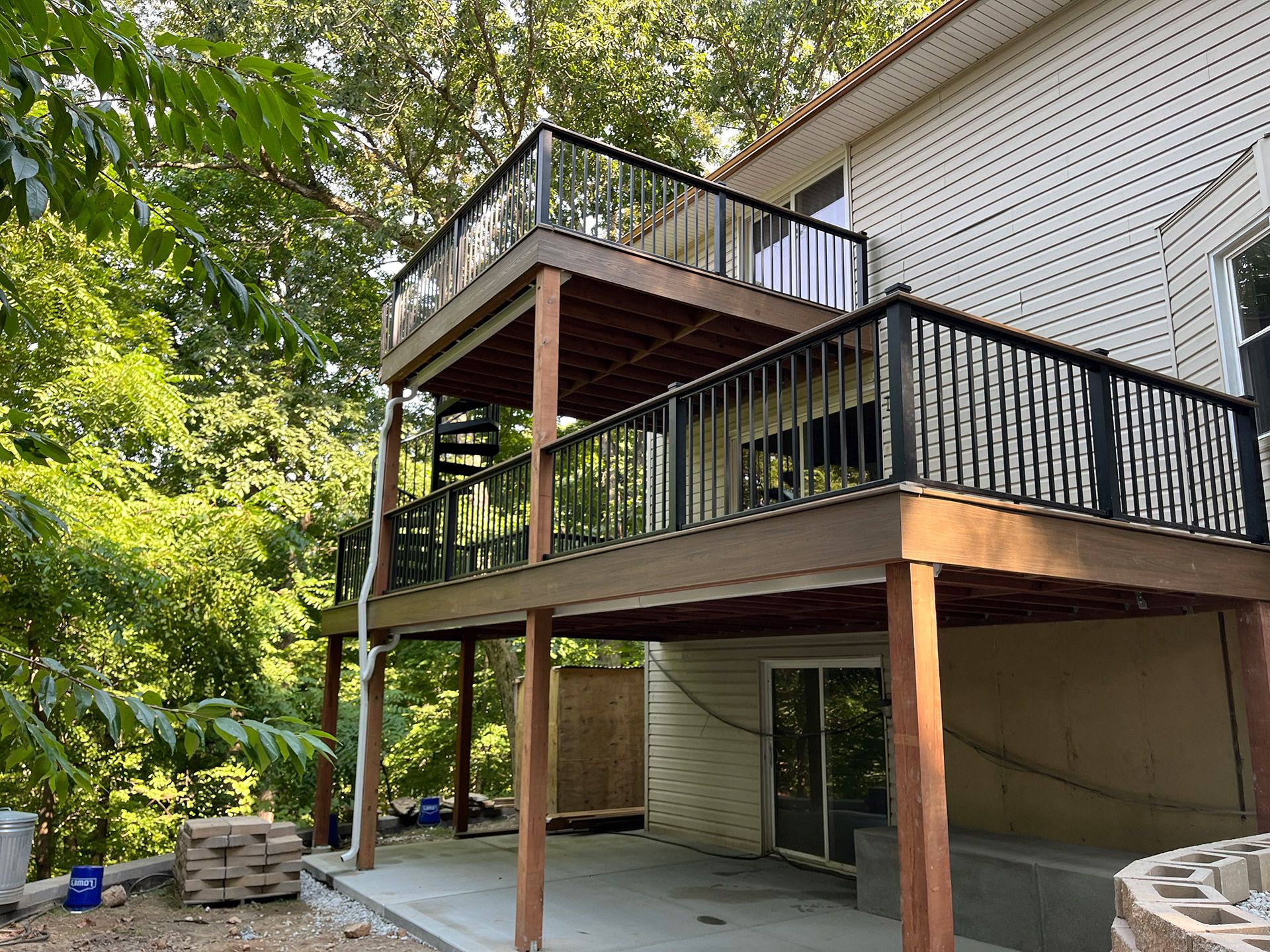 Back deck with black railings on a beige house, surrounded by trees and a patio below.