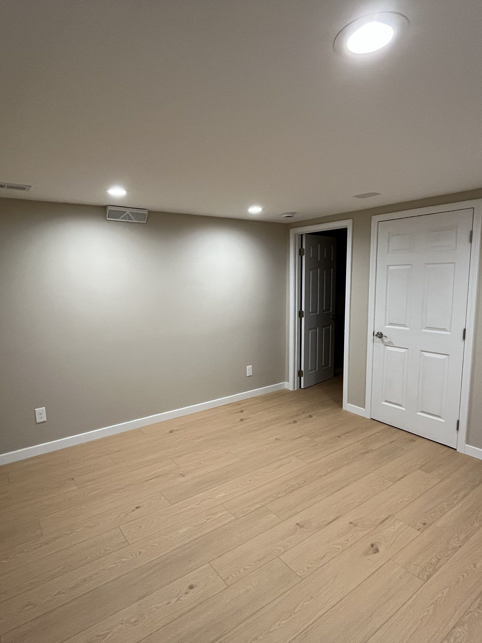 Empty beige bedroom with wood flooring, recessed ceiling lights, and white closet doors.