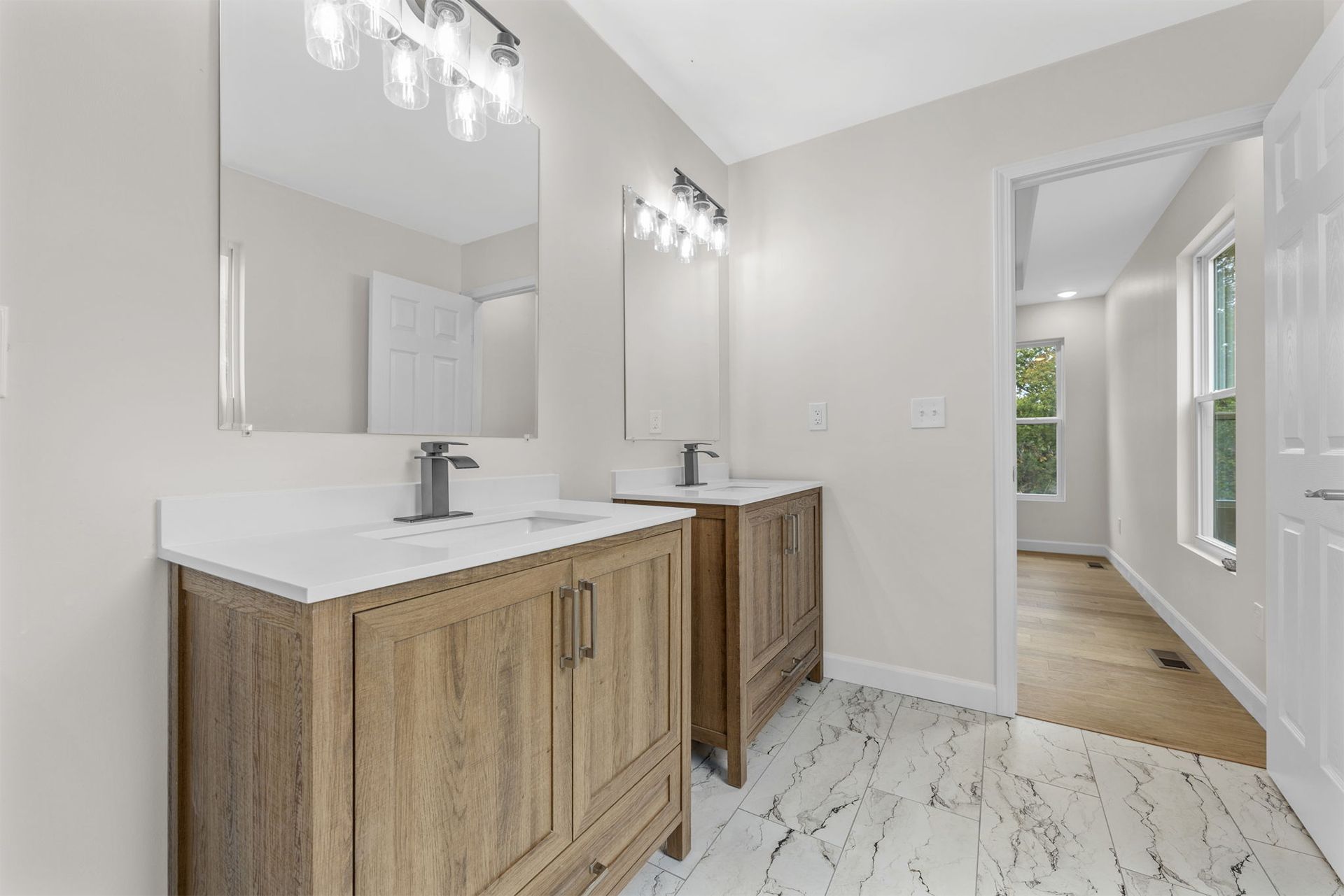 Bright bathroom with wood vanities, double sinks, large mirrors, and white marble-style tile flooring