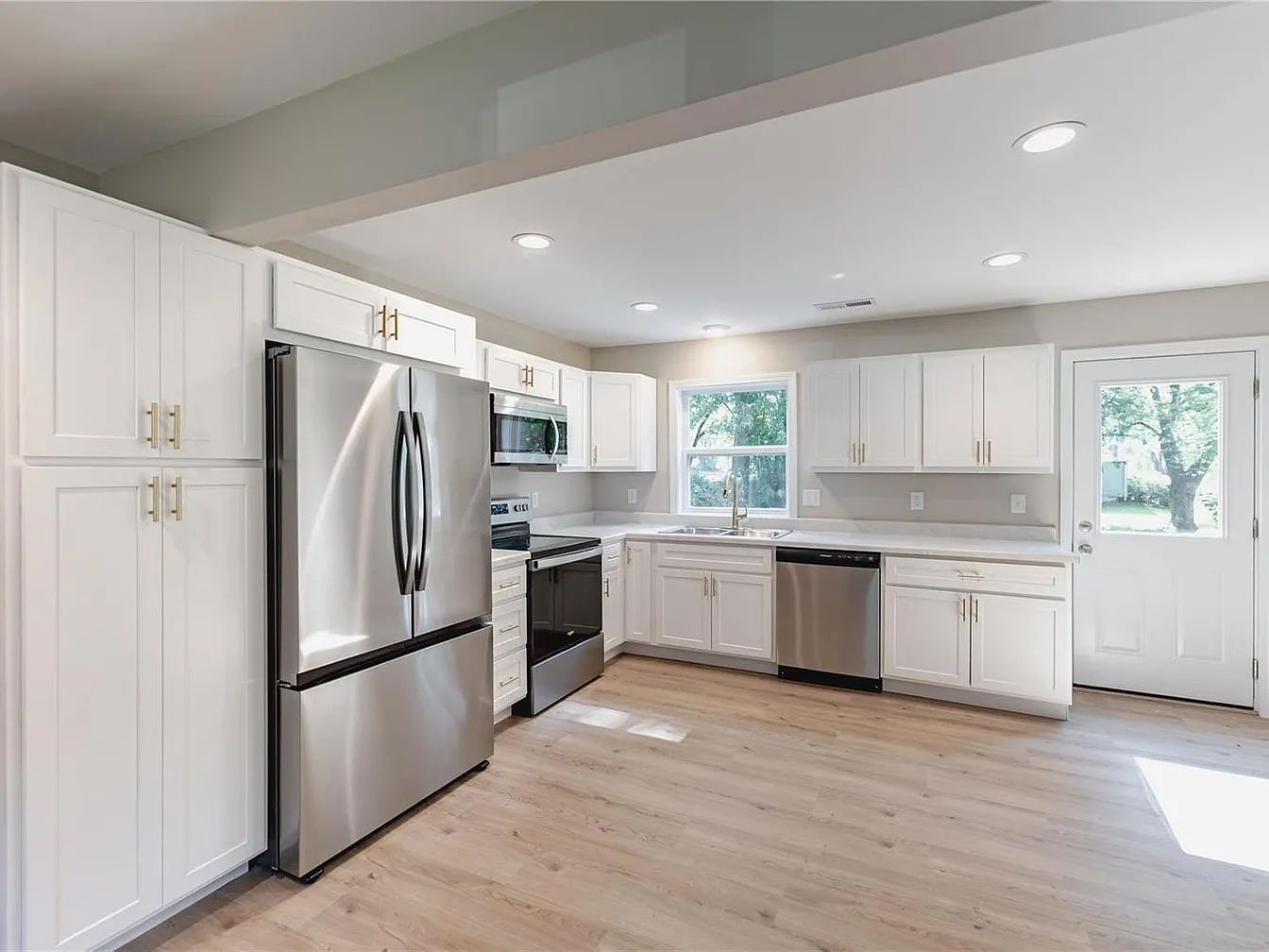 Bright white kitchen with stainless steel appliances, light wood floors, and large windows