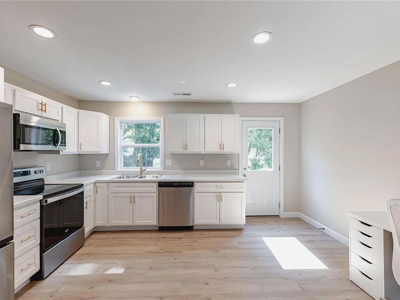 Bright white modern kitchen with stainless appliances, white cabinets, and sunlight on light wood floors