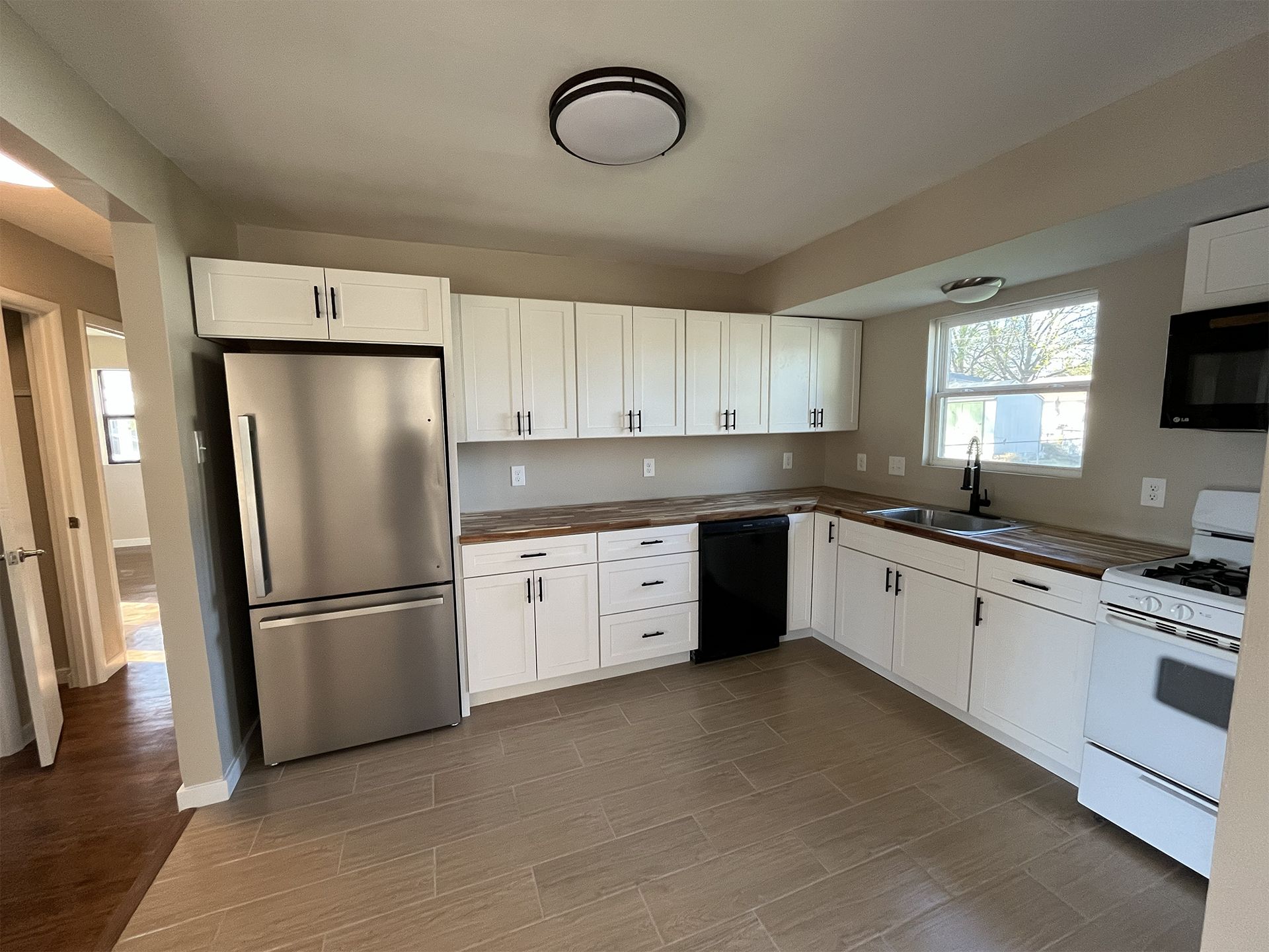 Bright kitchen with white cabinets, stainless fridge, tan tile floor, and a window over the sink.