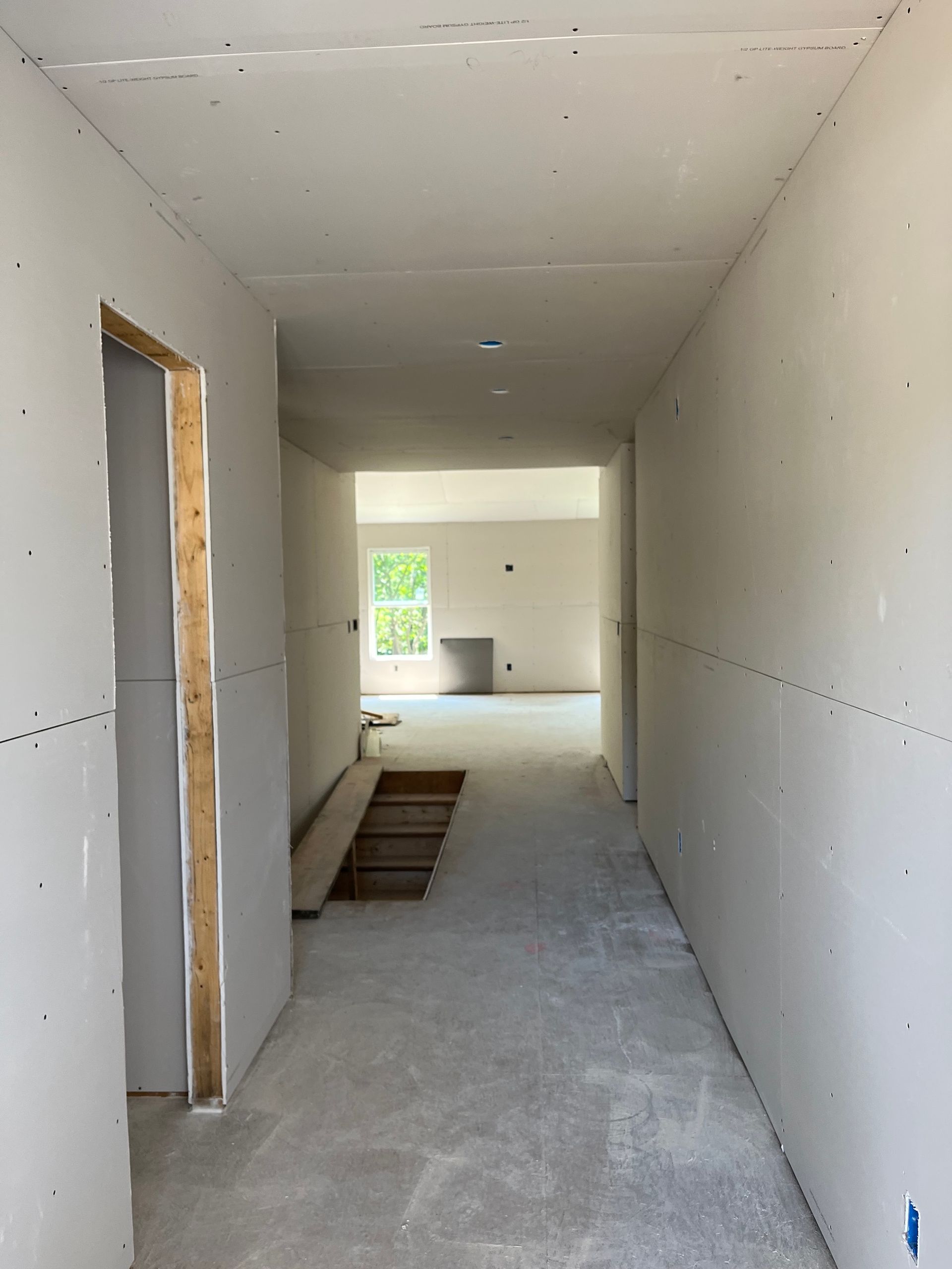 Unfinished hallway with white drywall, a doorway on the left, and a small step up to a bright room ahead