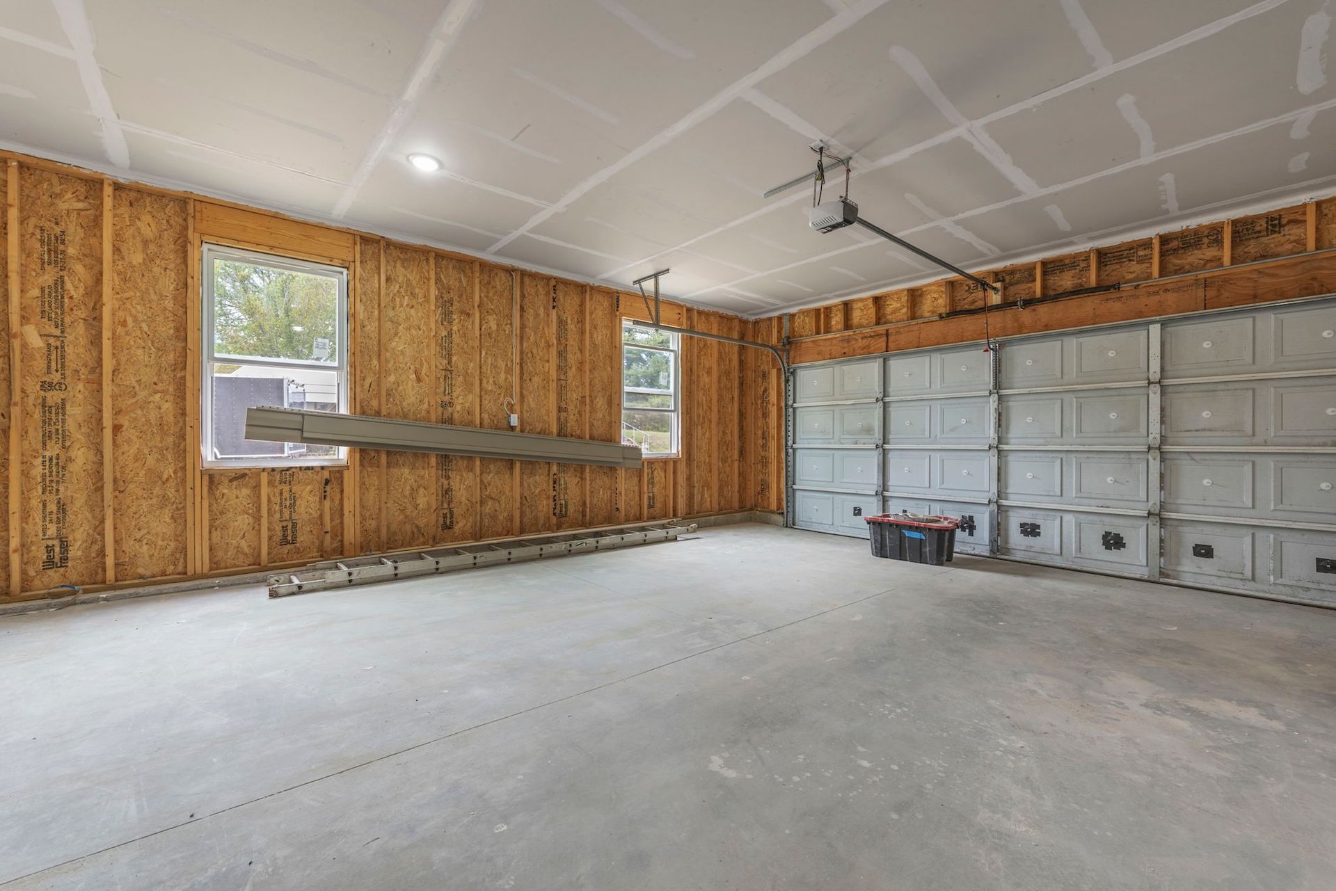 Empty unfinished garage with bare plywood walls, concrete floor, windows, and a closed white garage door.
