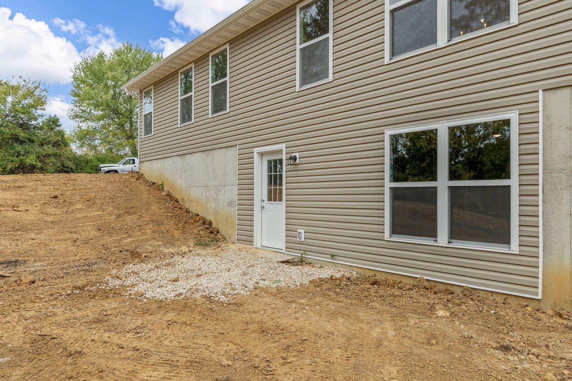 Rear view of a beige two-story house with a small side door, windows, and a dirt yard under construction.