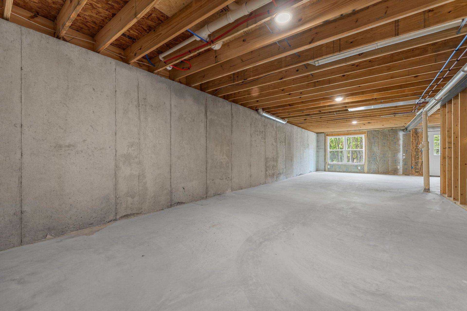 Unfinished basement with concrete walls, exposed wood ceiling joists, and a small window at the far end.