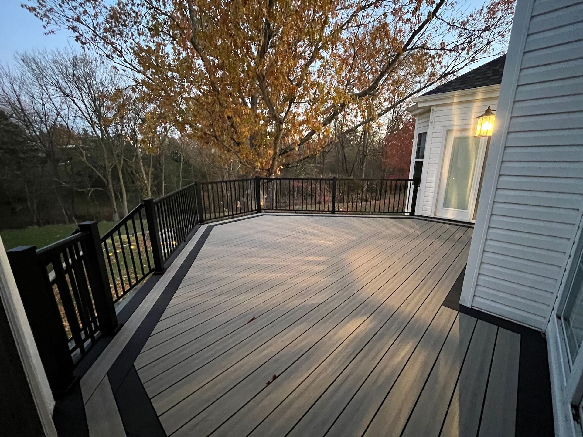 Curved wooden deck with black railing beside a blue house, overlooking autumn trees.