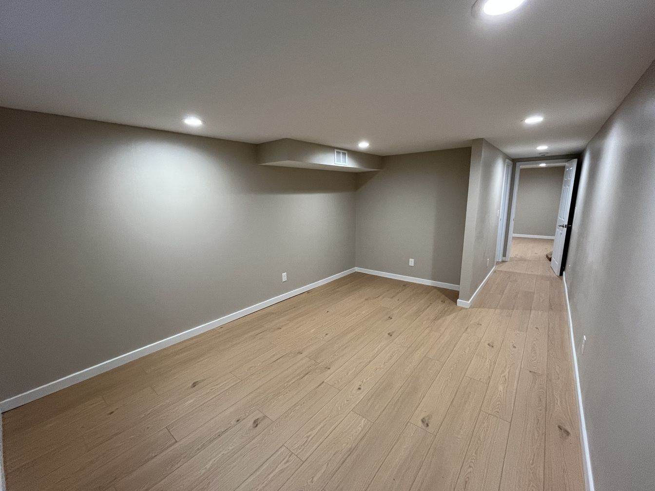 Empty finished basement room with light wood floors, gray walls, recessed lights, and a small window.