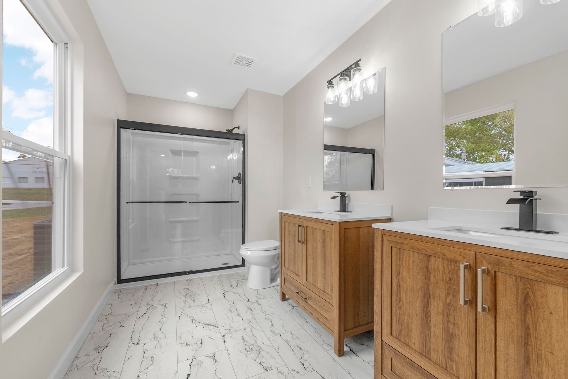 Bright bathroom with wood vanity, black-framed shower, white tile floor, and large window