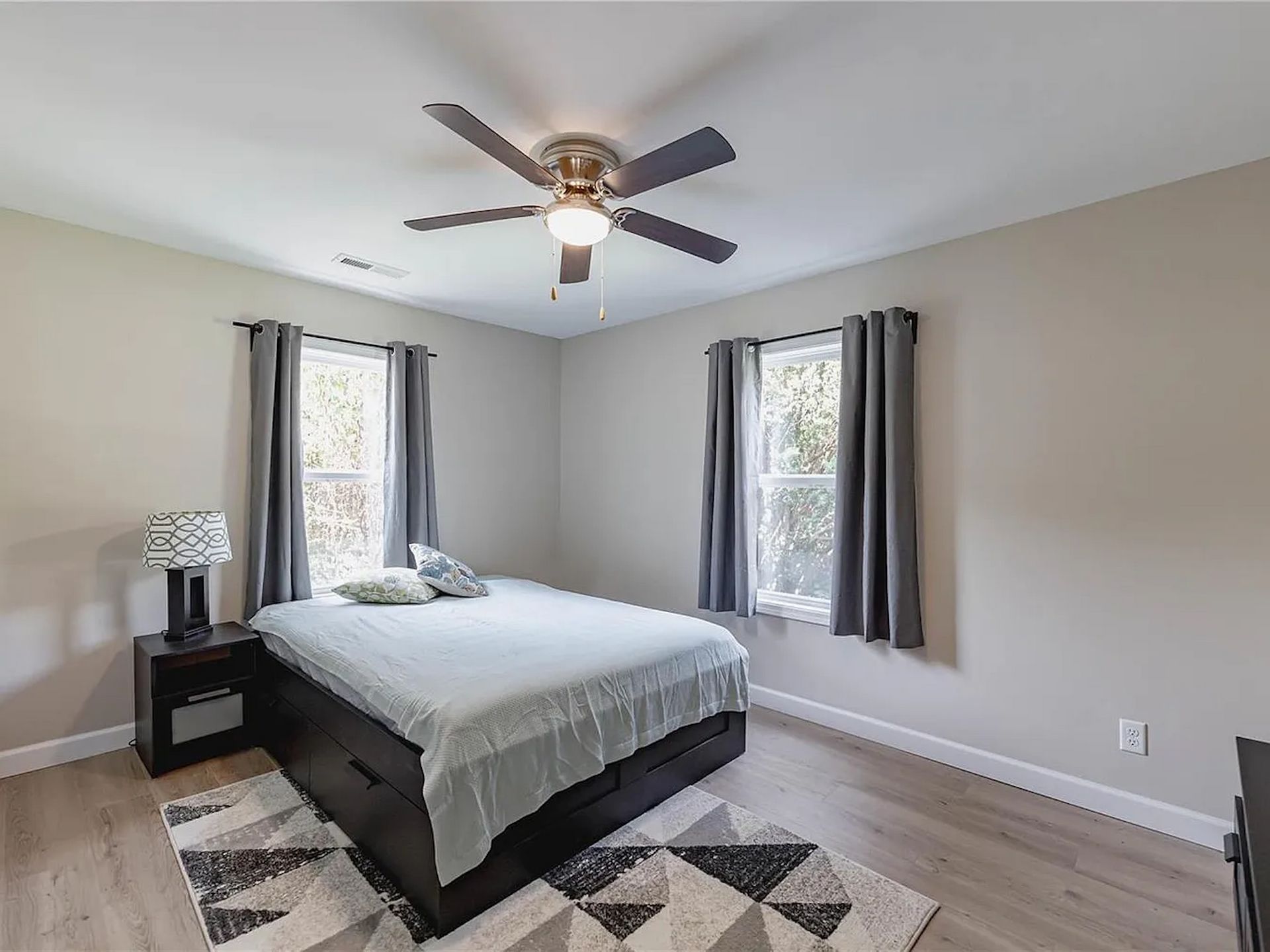 Bright bedroom with a bed, bedside table, ceiling fan, gray curtains, and black-and-white rug.