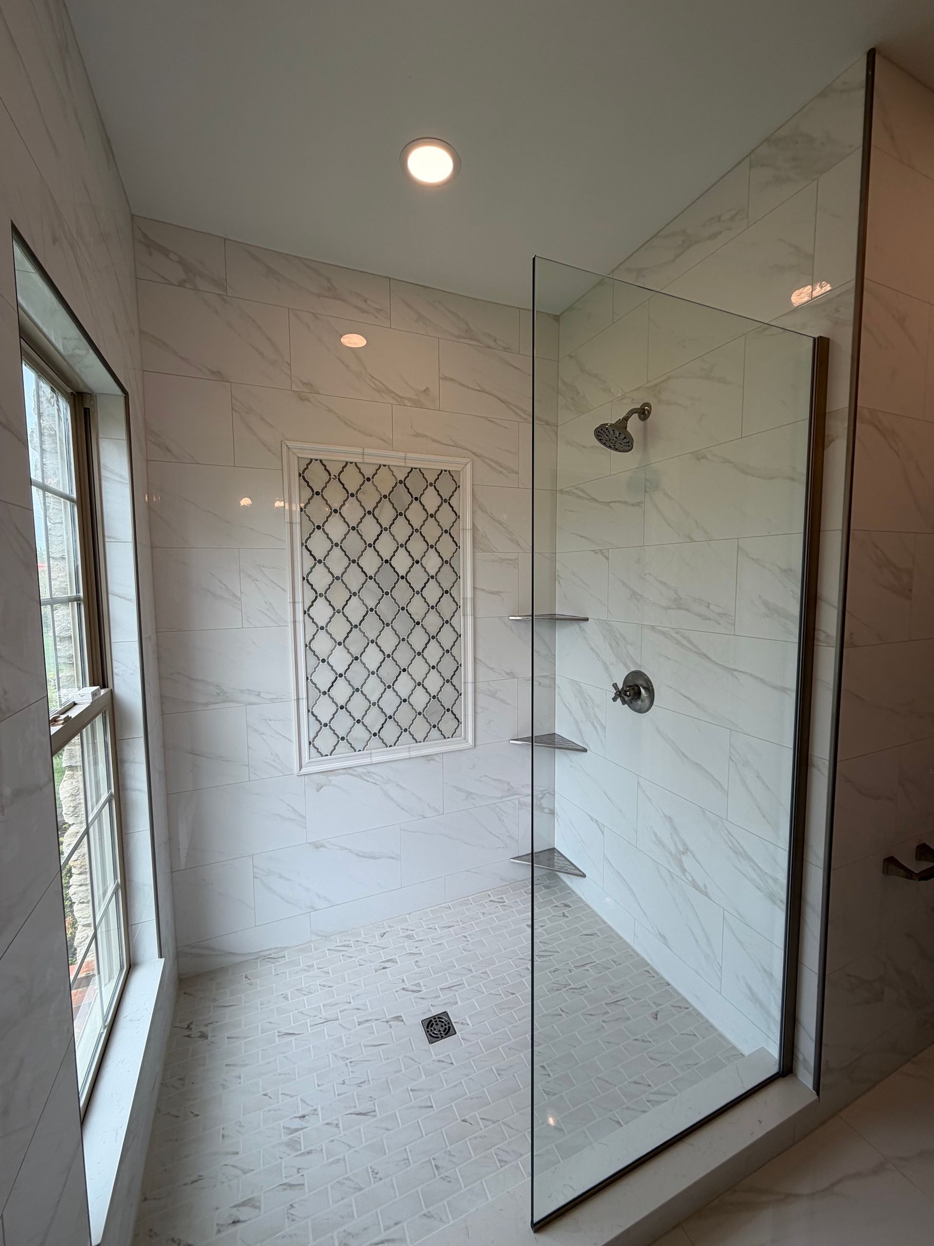 Modern shower stall with white tile, glass door, built-in shelves, and a window nearby