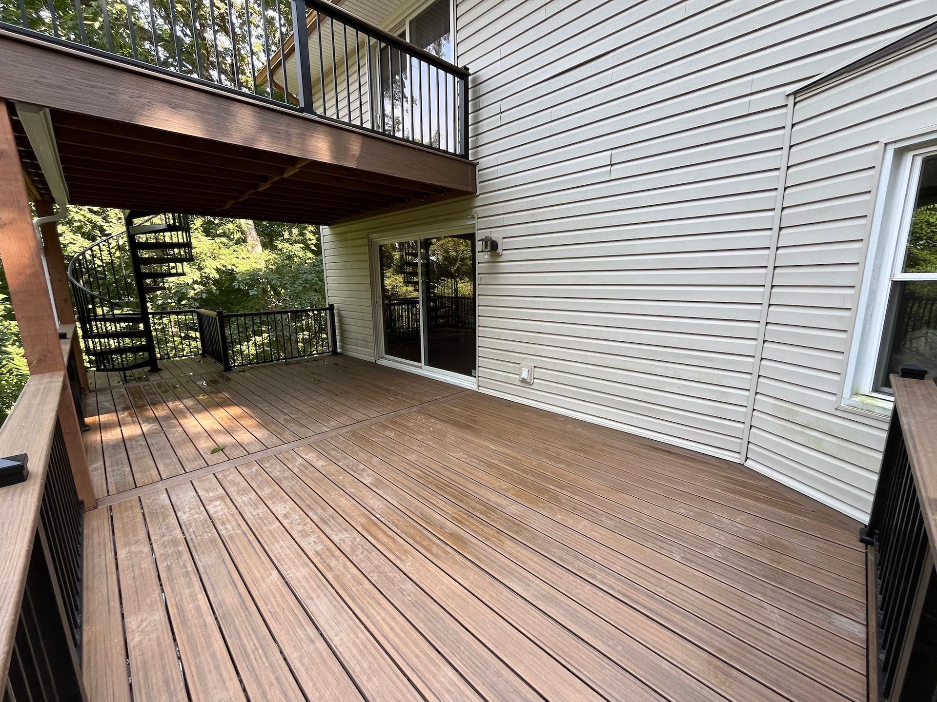 Wooden deck beside a beige house, with upper balcony and black railings overlooking a wooded yard.