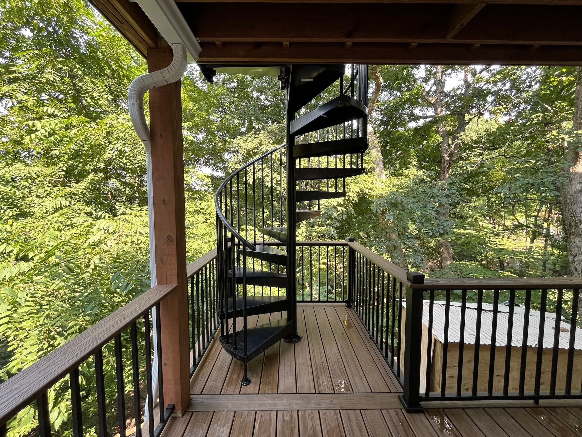 Covered wooden porch with black railings and a spiral staircase overlooking trees