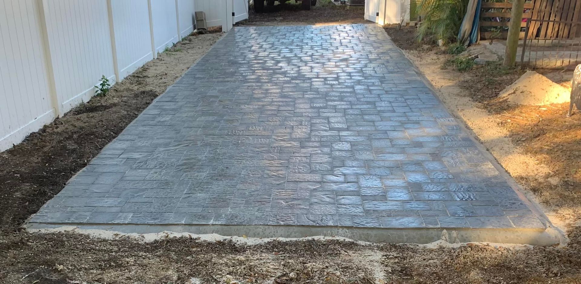 Concrete driveway with a brick-like pattern. White fence on the left, dirt on the sides, and a vehicle in the distance.