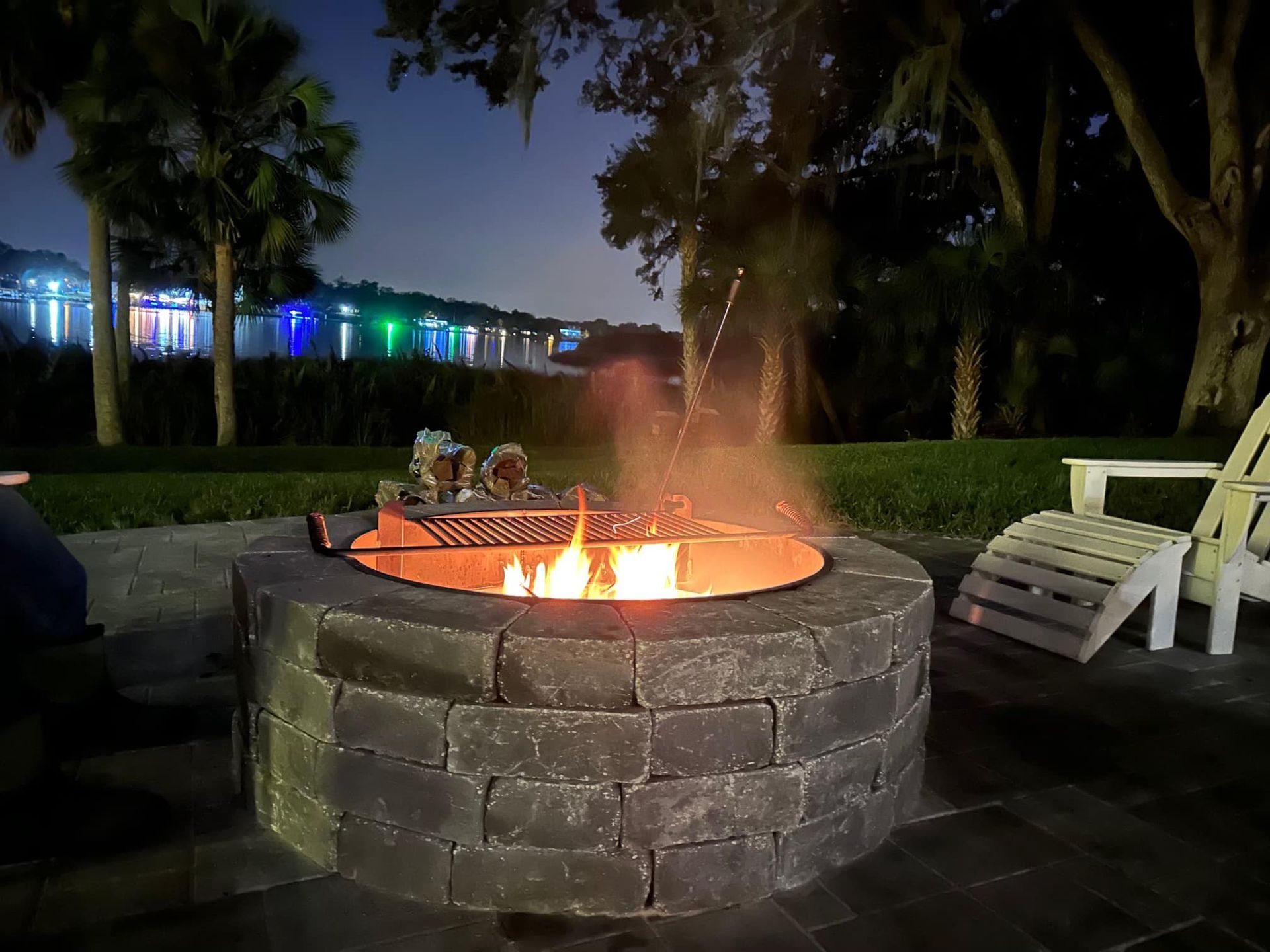 Fire pit with a burning fire at night. Two white chairs nearby. Water and lights in the background.
