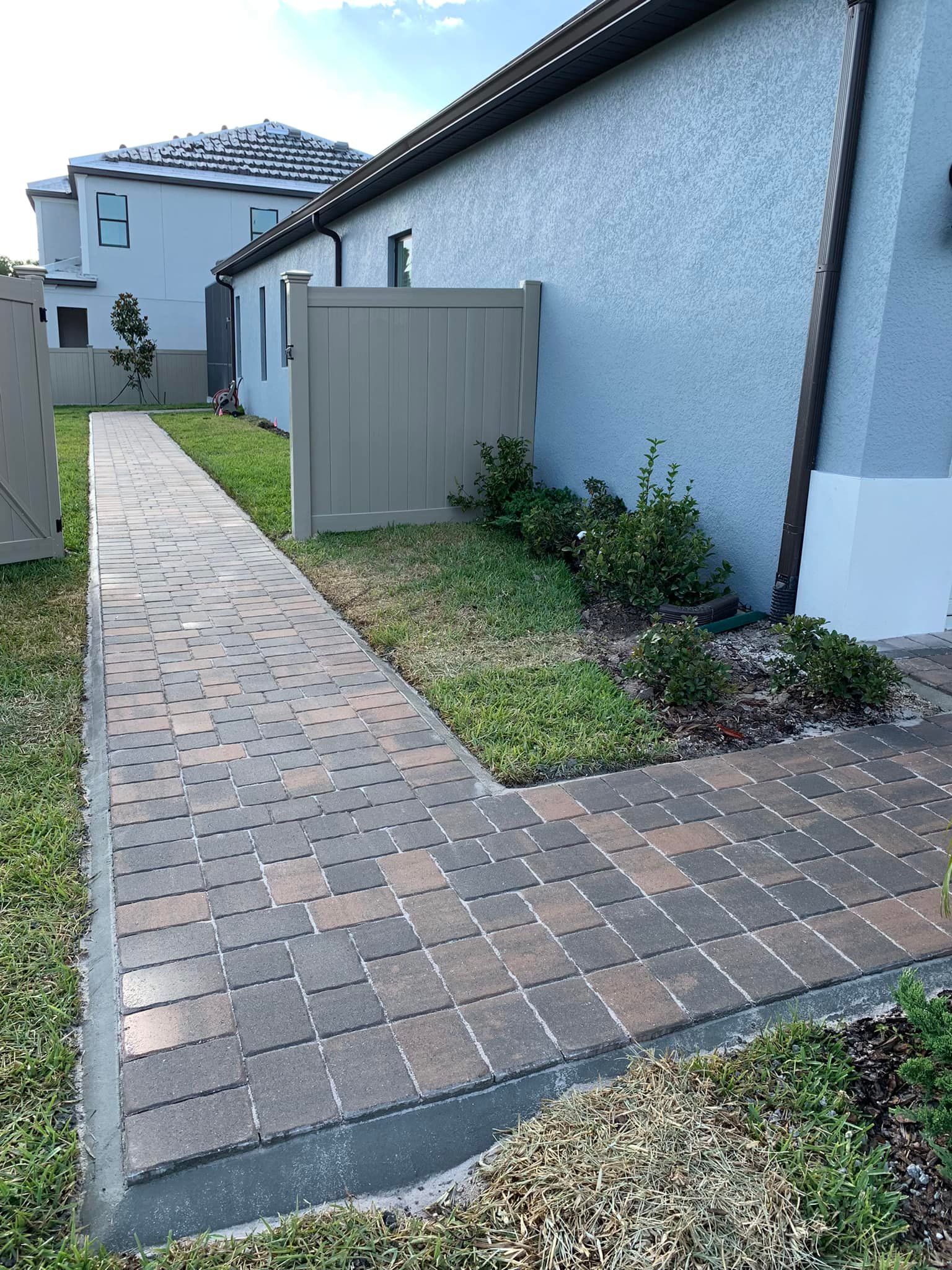 Brick walkway next to a house with a tan fence and small shrubs.