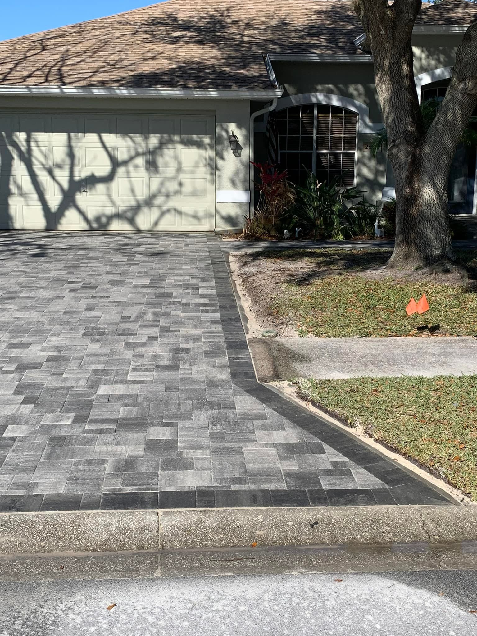 Paver driveway in front of a house with a gray garage door. A tree shades the right side.