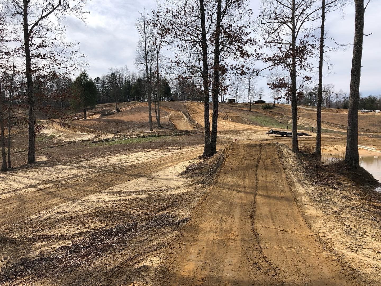 A dirt path leads through a cleared, wooded area with exposed soil and scattered trees under a cloudy sky.