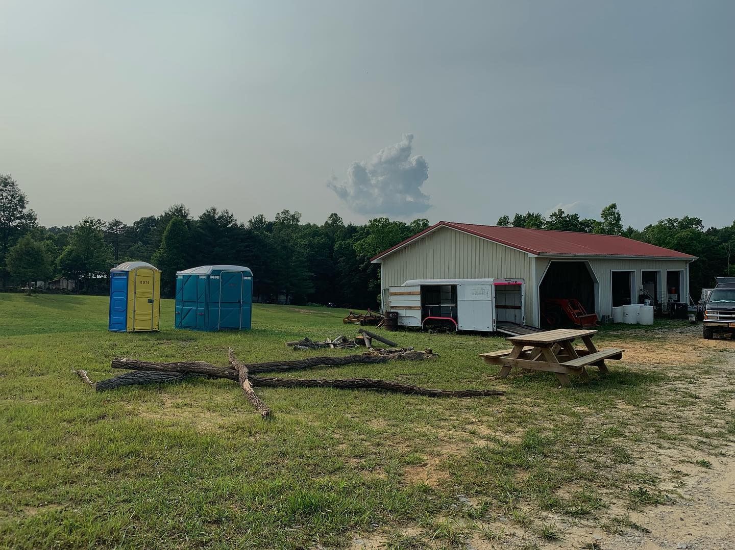 A grassy field with two portable toilets, fallen logs, a picnic table, and a white building with a red roof.
