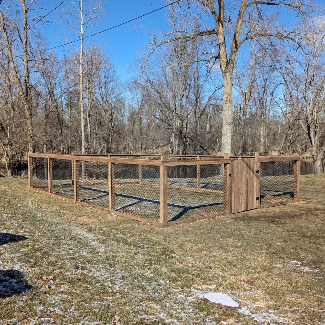 A rectangular garden enclosure with wooden posts and wire fencing, including a gate, set in a grassy yard near trees. A rectangular garden enclosure with wooden posts and wire fencing, including a gate, set in a grassy yard near trees.
