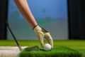 Hand in golf glove placing a white golf ball on green artificial turf next to a club.