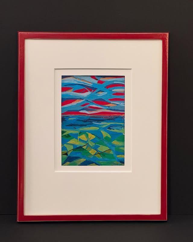A landscape painting of a blue sea and vibrant, layered sky, housed in a white mat and a bright red frame.