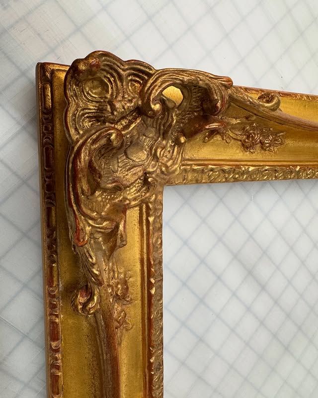 A close-up of a gold ornate picture frame corner with carved, scrolling decorative patterns against a grid background.
