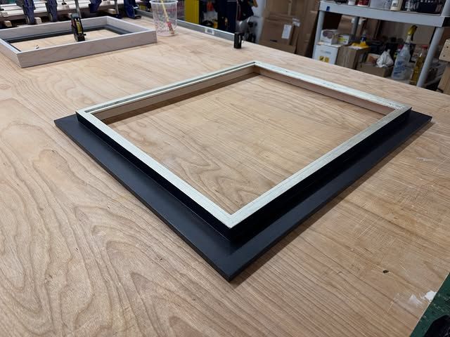 A metal picture frame sits centered on a black mounting board, resting on a wooden workshop table.